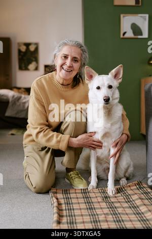 Porträt einer älteren kaukasischen Frau, die lächelt, während sie einen Hund im Haus streichelt Stockfoto