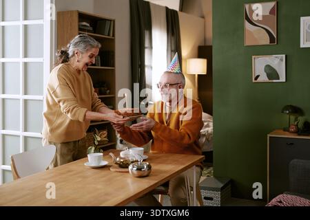 Ältere kaukasische Frau schenkt dem älteren kaukasischen Mann lächelnd Geburtstagskuchen Stockfoto