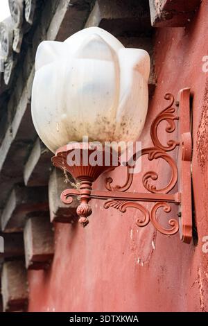 Nahaufnahme einer Vintage-Wandleuchte im Freien mit kunstvoller roter Metallhalterung und mattiertem weißem Glaskugel auf verwittertem rotem Stuckgebäude, Architectural de Stockfoto