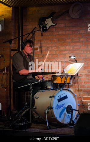 Schlagzeuger, der ein Set in einem gemütlichen, mit Backsteinmauern umgebenen Musikort spielt, beleuchtete Noten und warme Bühnenbeleuchtung, elektrische Gitarre an der Wand. Stockfoto