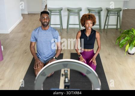 Mann und Afroamerikanerin in Sportkleidung kniend auf Matten zu Hause, filmen mit Ringlicht Stockfoto