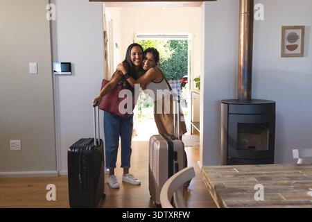 Verschiedene Freundinnen umarmen sich und halten Koffer auf Rädern beim Hauseingang mit Holzofen Stockfoto