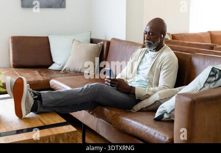 Reifer afroamerikanischer Mann, der zu Hause auf dem Sofa liegt und das Telefon hält, Ohrstöpsel trägt, Füße auf dem Tisch Stockfoto