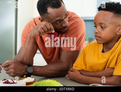 Afroamerikanischer Vater und Sohn im Schulalter bereiten Salat in der Küche vor, mit gekräuseltem Pfeffer Stockfoto