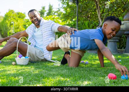 Afroamerikaner Vater und Sohn in blauem Polo kniend auf dem Rasen und sammeln Eier in weißen Korb Stockfoto