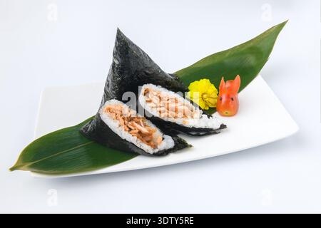 Traditionelle japanische Onigiri-Reiskugel gefüllt mit gekochtem Lachs, in Algen gewickelt und auf einem Bambusblatt mit dekorativem Gemüse serviert. Stockfoto