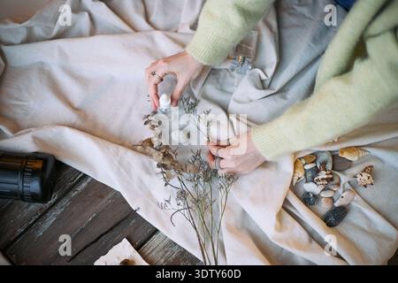 Kaukasische Frau, die getrocknete Blumen und dekorative Steine auf Stoffoberfläche für Produktfotografie anordnet, Hände sichtbar halten Glasflasche, natürliche Elemente um die Komposition verteilt Stockfoto