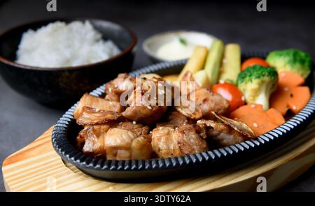 Japanische Schweinesteakwürfel, serviert auf einer heißen Platte, begleitet von gedämpftem Reis, cremiger Suppe und verschiedenen Gemüsesorten in rustikalem Stil Stockfoto
