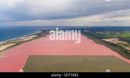Aus der Vogelperspektive auf einen großen rosafarbenen Salzsee, der von einer Sandküste und dem offenen Meer unter einem bewölkten Himmel begrenzt wird. Stockfoto
