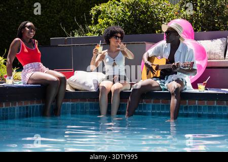 Drei Freunde sitzen am Pool im Hinterhof, mit Füßen im Wasser, ein Mann mit Cocktail, der Gitarre spielt Stockfoto