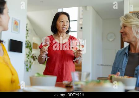 Verschiedene Freundinnen hören zu, während eine ältere Frau in roter Bluse zwei Weingläser am Tisch hält Stockfoto