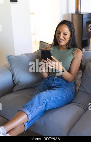 Afroamerikanerin, die auf einem grauen Sofa sitzt und ihr Smartphone mit Olive-Top und Jeans hält Stockfoto