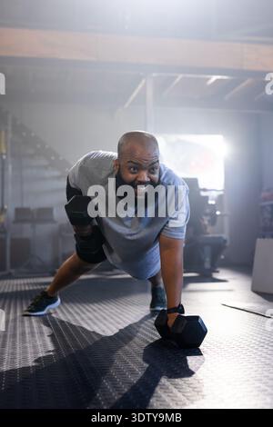 Mann mit Hexenhantel rudert in einer Planke auf strukturiertem Gummiboden und trägt graues Hemd Shorts Tracker Stockfoto