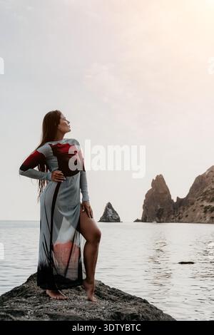 Frau, Ozean, Klippen - Eine Frau in einem langen Kleid steht auf einem Felsen mit Blick auf das Meer und die Klippen. Stockfoto