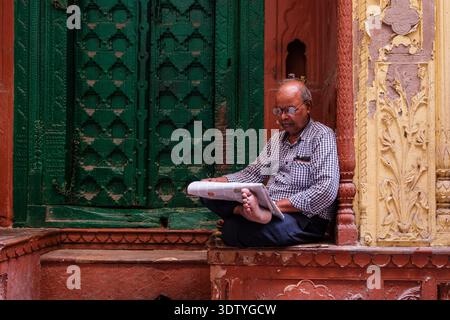 Ein Mann sitzt auf einem dekorativen Steinvorsprung vor einer verzierten grünen Tür in einer engen Gasse in Alt-Delhi. Stockfoto
