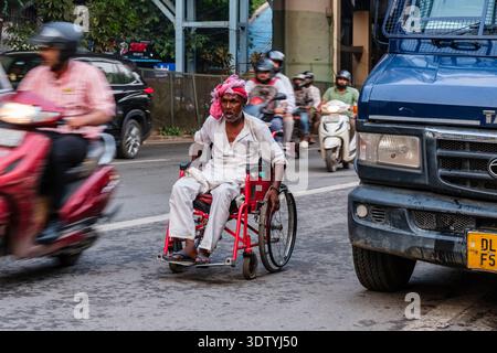 Ein Mann im Rollstuhl navigiert durch den belebten Verkehr in Neu-Delhi, umgeben von fahrenden Autos und Motorrädern mit einer Brücke in der Ferne. Stockfoto