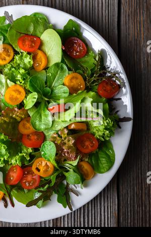 Gemischter Salat mit Kirschtomaten auf weißem Teller. Frisches Blattgemüse auf hölzernem Hintergrund Stockfoto