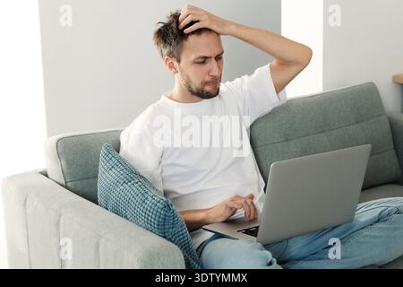 Frustrierter Millennial-Geschäftsmann, der sich nach einem Geschäftsausfall deprimiert fühlt, schauen Sie sich den Laptop-Monitor an, während Berichte über Finanzbankrott weinen. Gelegenheitsmann liest E-Mail mit schlechten Nachrichten Sitzen Sie auf der Couch zu Hause || Model veröffentlicht Stockfoto