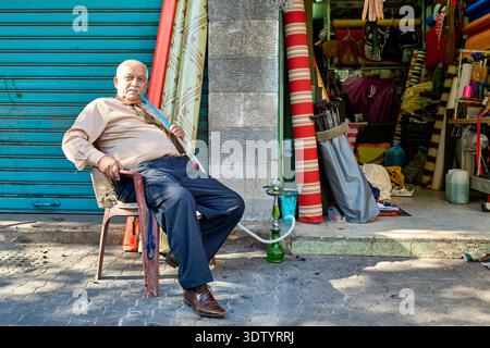 Ein älterer Mann sitzt auf einem Stuhl in Amman. Er raucht gerne Shisha, während er sich neben seinem Laden ausruht. Die Straße zeigt lokale Farben und Elemente an. Stockfoto