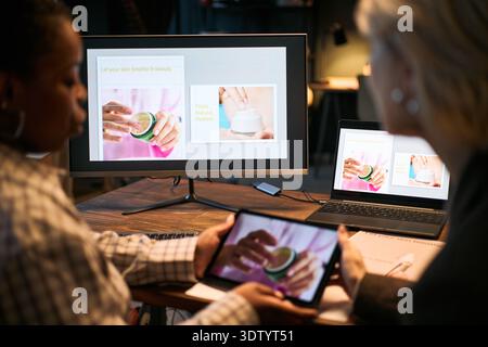 Zwei erwachsene Frauen, eine Schwarze und eine Kaukasierin, arbeiten an der Präsentation von Hautpflegeprodukten mit einem digitalen Tablet und einem Computermonitor mit kosmetischen Bildern zusammen Stockfoto