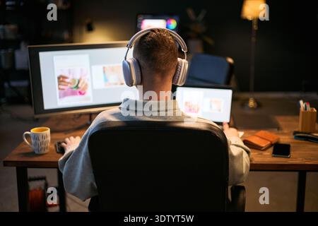 Kaukasischer junger Erwachsener, der am Schreibtisch sitzt, Computer und Laptop benutzt, während er Kopfhörer trägt, an einem digitalen Projekt mit Kaffeetasse und Notebook arbeitet, das am Arbeitsplatz sichtbar ist Stockfoto