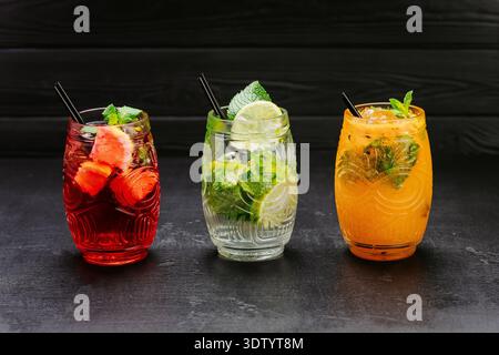 Farbenfrohe, frische Cocktails mit Minze, Limette und Obst, serviert in Gläsern auf dunklem Hintergrund Stockfoto