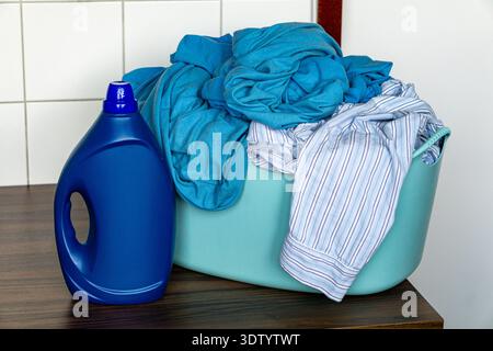 Wäschekorb aus Plastik gefüllt mit abgenutzter Kleidung auf Holzfläche neben der blauen Waschmittelflasche. Stockfoto