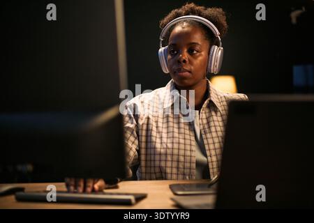 Schwarze junge Erwachsene Frau, die Kopfhörer trägt, arbeitet am Computermonitor in moderner Büroumgebung, fokussierter Ausdruck, mit Tastatur und Maus, digitaler Arbeitsbereich sichtbar Stockfoto