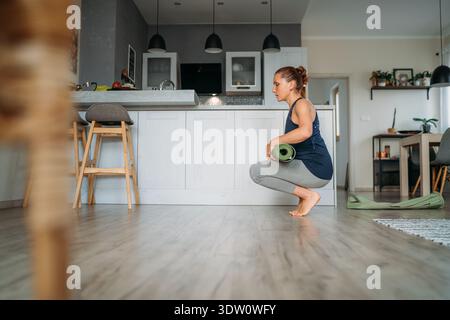 Gesunde junge Frau, die sich auf einem Holzboden hockt, während sie achtsam ihre grüne Yoga-Matte aufrollte, nachdem sie eine morgendliche Wellness-Routine in einem hellen Stockfoto