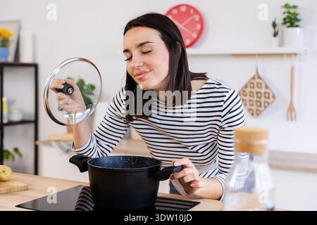 Junge glückliche Frau, die frisches Aroma von Suppe riecht, während sie in der Küche kocht Stockfoto