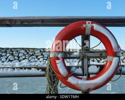 Roter Rettungsschirm bedeckt mit Schnee auf Holzgeländer vor gefrorenem Wasser unter blauem Winterhimmel. Kopierraum Stockfoto