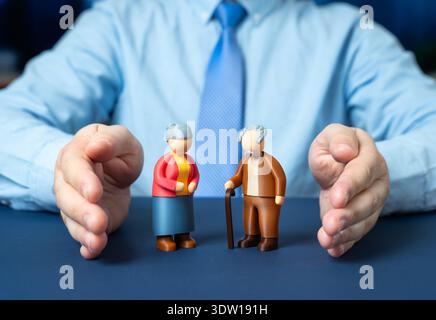 Ein Mann beschützt mit seinen Händen die Figuren von ein paar älteren Rentnern. Sozialschutz und finanzielle Sicherheit. Unterstützung für Senioren und pensi Stockfoto