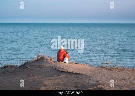 Eine Frau in einer orangefarbenen Jacke sitzt mit einem Hund auf einer Sanddüne und schaut auf die blaue Oberfläche des Kapchagai-Stausees. Die Landschaft vermittelt die Atmosphäre Stockfoto
