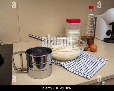Prozess der Herstellung von Teig in einer Mischschüssel auf einem Küchentisch. Schneebesen, Siebe, Mehl, Ölflaschen und Eier sind in der Nähe angeordnet. Flüssiger Pfannkuchenteig Stockfoto
