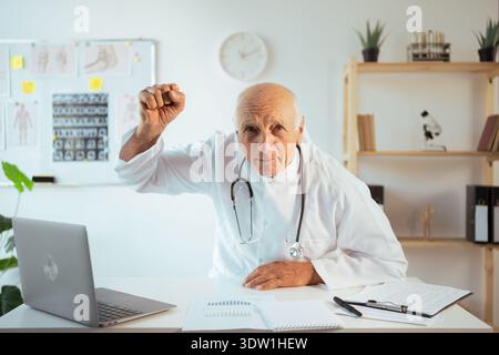 Wütender männlicher Senior Doctor Mann, der weißen Mantel Arbeit auf Laptop in der Krankenhausklinik trägt, schreiend und geballt die Faust Stockfoto