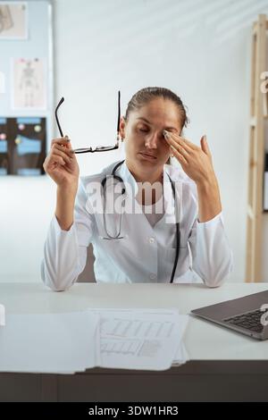 Überarbeitete junge Ärztin im Krankenhausbüro reibt ihre müden, trockenen Augen, hat Augenanstrengung durch ständige Arbeit auf dem Laptop Stockfoto