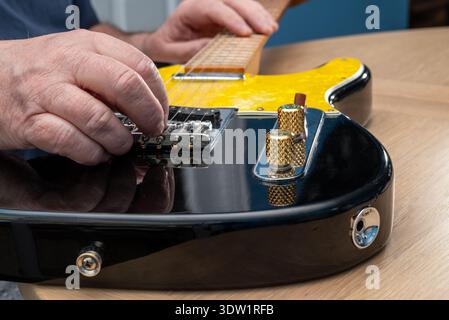 Die Hände einer Person werden gezeigt, wie sie die Saiten auf einer gelben und schwarzen E-Gitarre einstellen, wahrscheinlich stimmen oder vor dem Spielen Anpassungen vornehmen. Stockfoto