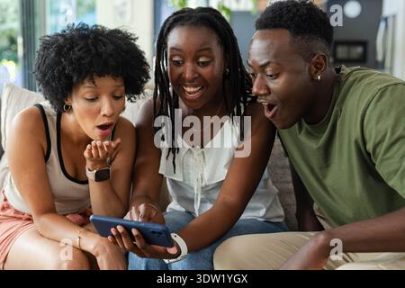 Afroamerikanische Freunde beugten sich nach vorne und keuchten auf einem hellen Sofa an einem Smartphone mit blauem Gehäuse Stockfoto