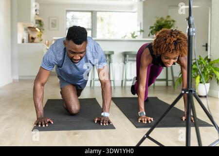 Diverse Paare, die Dielen auf Matten zu Hause mit Stativ halten und Fitnessuhren in Sportbekleidung tragen Stockfoto