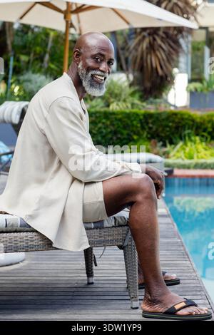 Ranghoher afrikanischer Mann, der auf einem gewebten Rattanstuhl am Poolrand sitzt und leichte beige Loungewear trägt Stockfoto