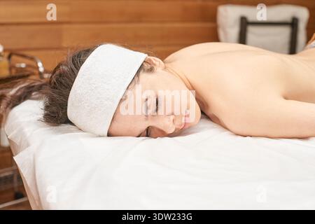 Entspannte Junge Kaukasische Frau Genießt Spa-Massagesitzung Auf Dem Tisch Mit Weißem Handtuch-Stirnband In Ruhiger Umgebung. Stockfoto