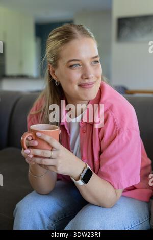 Frau, die mit beiden Händen einen pfirsichfarbenen Keramikbecher hält, während sie auf einem grauen Sofa sitzt und eine weiße Smartwatch trägt Stockfoto
