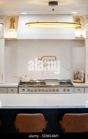 Moderne cremefarbene Kücheneinrichtung mit Marmor Backsplash, Gasherd, Messingtopf Füllung und Pendelbeleuchtung in einem sauberen, modernen Zuhause. Stockfoto