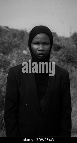 Schwarz-weißes Porträt einer jungen Afrikanerin, die einen Hijab und einen formellen schwarzen Anzug trägt und im Freien mit einem ruhigen, selbstbewussten Ausdruck steht. Stockfoto