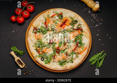 Gourmet-Pizza mit Lachs-Rucola-Käse und sonnengetrockneten Tomaten auf Holzbrett Stockfoto