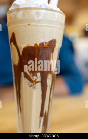 Ein hohes Glas gefüllt mit einem cremigen Milchshake, garniert mit Schlagsahne und Schokoladensoße in einem gemütlichen Café. Stockfoto