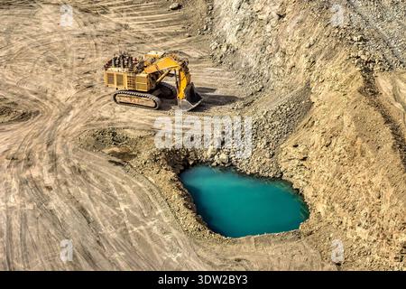 Hochwinkelansicht einer großen gelben hydraulischen Bergbauschaufel, die auf felsigem Gelände in einem Tagebau arbeitet, mit einem türkisfarbenen Wasserbecken im for Stockfoto