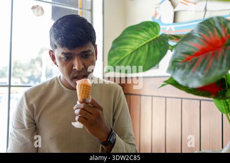 Ein junger Mann sitzt in einem Café und isst eine Eiscreme, während er nachdenklich aussieht. Stockfoto