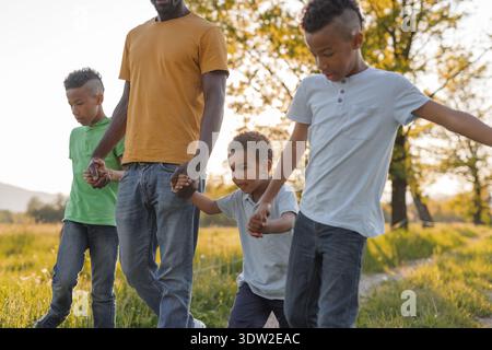 Ein Vater spaziert mit seinen drei Kindern, die Hände halten, während er die Natur bei einem warmen Sonnenuntergang genießt. Sie sind glücklich und verspielt zusammen. Stockfoto