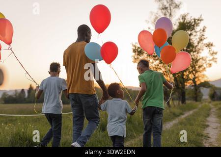 Alleinerziehender Vater genießt einen Tag im Freien mit seinen Kindern, während sie während des Sonnenuntergangs bunte Ballons halten, während sie einen Pfad hinunterlaufen. Stockfoto